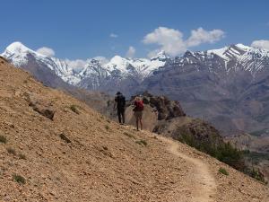 Two trekkers walking along a mountain trail in Spiti Valley with snow-covered Himalayan peaks in the background